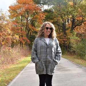 May include: A woman wearing a gray plaid coat, white shirt, and black pants stands on a paved path. The background features trees with autumn foliage in shades of orange, yellow, and green. Sunglasses are worn.