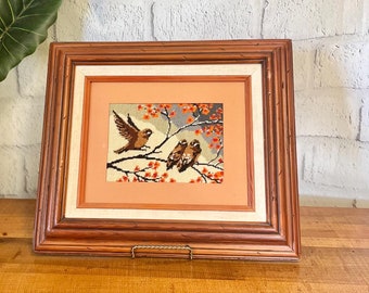 May include: A framed needlepoint artwork depicting three brown birds perched on a branch with orange blossoms. The frame is brown wood with a gold-colored metal stand.