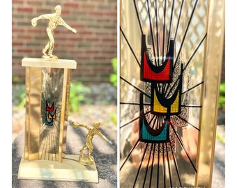 May include: A gold-colored bowling trophy with a stylized image of a bowler and three bowling pins. The trophy has a clear glass panel with a gold-colored background and a black and white image of a bowling lane. The trophy is on a wooden base with a gold-colored plaque that says "MADE WITH PICJOINTER".