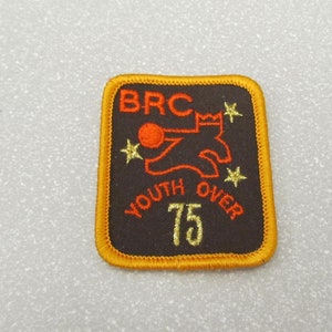 Puede incluir: Un parche bordado en negro y dorado con el texto "BRC Youth Over 75" y una ilustración roja y naranja de una persona lanzando una pelota.