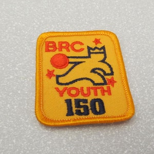 Puede incluir: Parche bordado amarillo con una ilustración en negro y rojo de una persona sosteniendo una pelota. El texto "BRC YOUTH 150" está bordado en negro.