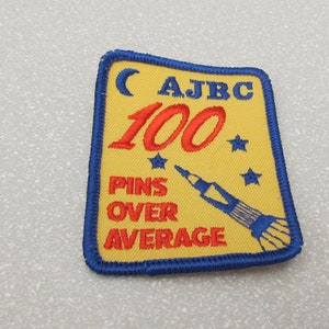 Puede incluir: Un parche bordado azul y amarillo con el texto "AJBC 100 PINS OVER AVERAGE", una luna creciente y estrellas. También se borda un cohete en el parche.