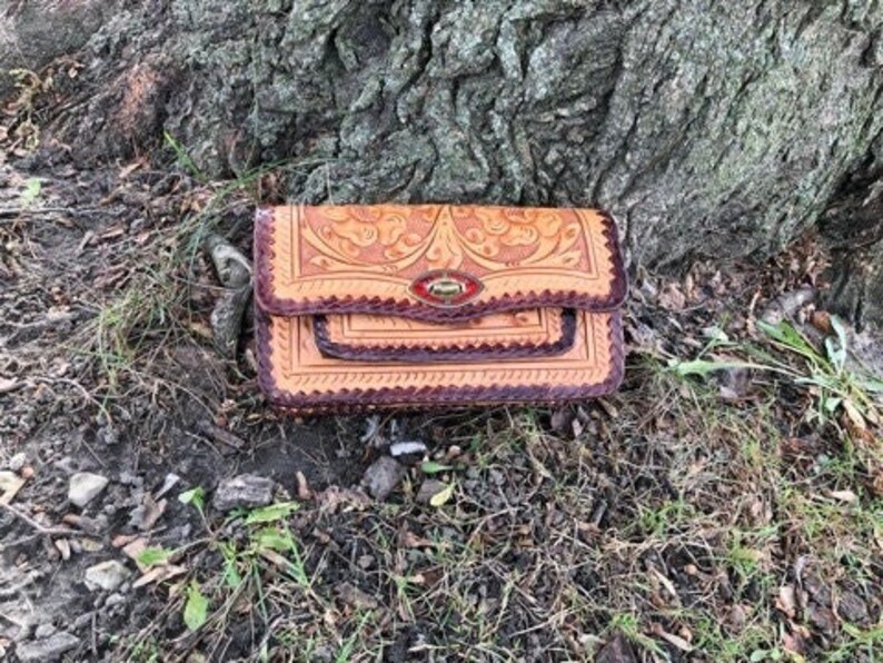 May include: A tan leather wallet with a floral embossed design and a dark brown border. The wallet has a flap closure with a red and gold clasp. The wallet is sitting on the ground near a tree.