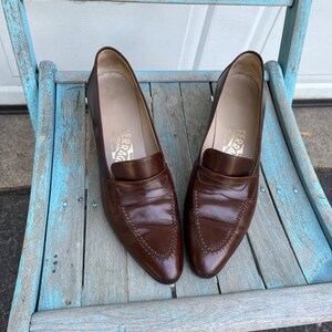 May include: A pair of brown leather loafers with a pointed toe and a stitched design. The loafers are sitting on a light blue wooden chair.