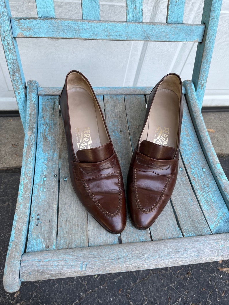May include: A pair of brown leather loafers with a pointed toe and a stitched design. The loafers are sitting on a light blue wooden chair.
