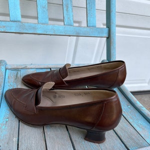 May include: A pair of brown leather heeled loafers with a pointed toe. The shoes have a small heel and a buckle detail on the top. The shoes are sitting on a light blue wooden chair.