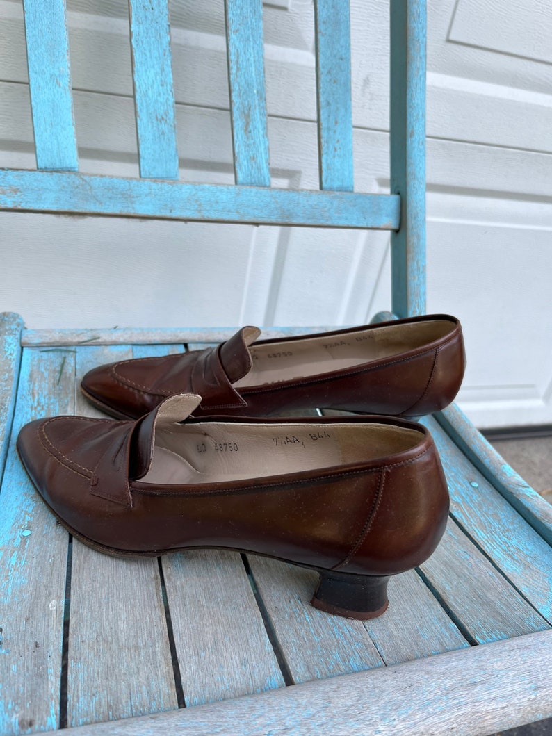 May include: A pair of brown leather heeled loafers with a pointed toe. The shoes have a small heel and a buckle detail on the top. The shoes are sitting on a light blue wooden chair.