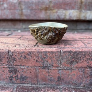 May include: A small, ceramic bowl with a textured, brown glaze. The bowl has a slightly irregular shape and is sitting on a brick surface.