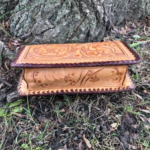 May include: A rectangular, light brown leather box with a dark brown braided border. The top and sides feature floral and leaf carvings. The box is resting on a bed of grass and leaves near a tree trunk.