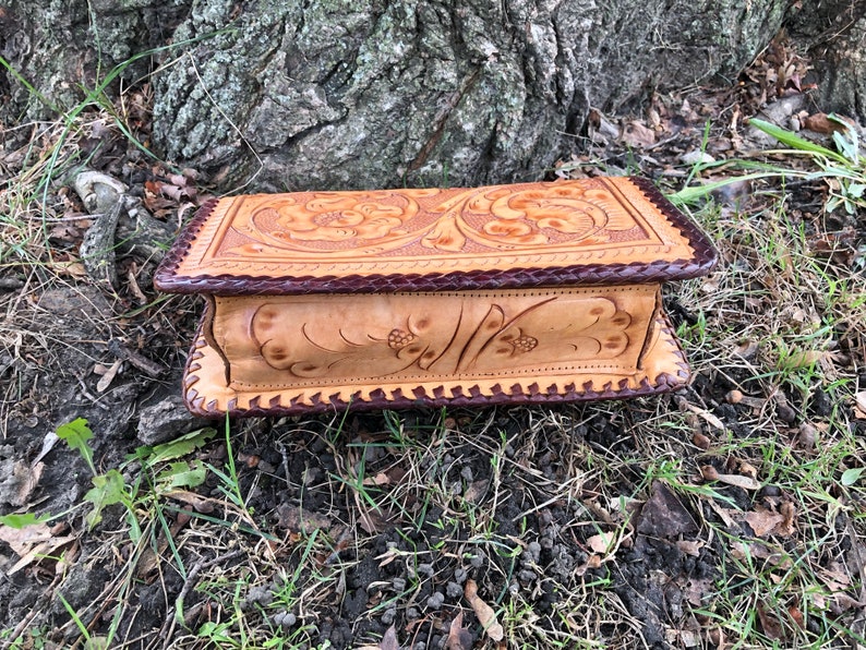May include: A rectangular, light brown leather box with a dark brown braided border. The top and sides feature floral and leaf carvings. The box is resting on a bed of grass and leaves near a tree trunk.