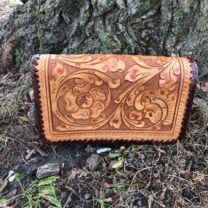 May include: A handcrafted leather clutch with intricate floral and scrollwork carving. The tan-colored leather is accented with a dark brown border and a scalloped edge. The clutch is set against a natural outdoor backdrop.