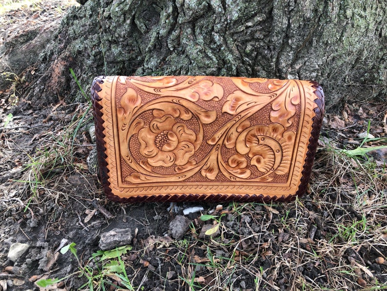 May include: A handcrafted leather clutch with intricate floral and scrollwork carving. The tan-colored leather is accented with a dark brown border and a scalloped edge. The clutch is set against a natural outdoor backdrop.