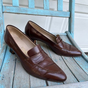 May include: A pair of brown leather heeled loafers with a pointed toe. The loafers have a gold-colored logo on the side that reads "Ferragamo".