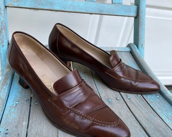 May include: A pair of brown leather heeled loafers with a pointed toe. The loafers have a gold-colored logo on the side that reads "Ferragamo".