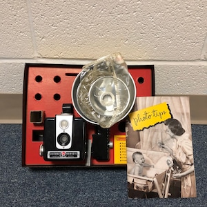 May include: Vintage Brownie Hawkeye Camera Flash Model kit in a red box. The kit includes a black camera, flash attachment, and a photo tips card with a black and white image of a mother and baby. The camera has the words "Brownie Hawkeye Camera Flash Model" printed on it.