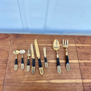 May include: Set of eight gold-tone silverware with black handles. The set includes a knife, fork, spoon, and four smaller spoons. The handles are decorated with intricate designs.