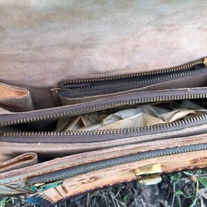 May include: Open view of a vintage leather satchel. The bag is tan with a detailed embossed design and multiple zippered compartments. The zippers and clasp are gold-toned. The interior is lined with a lighter tan material.