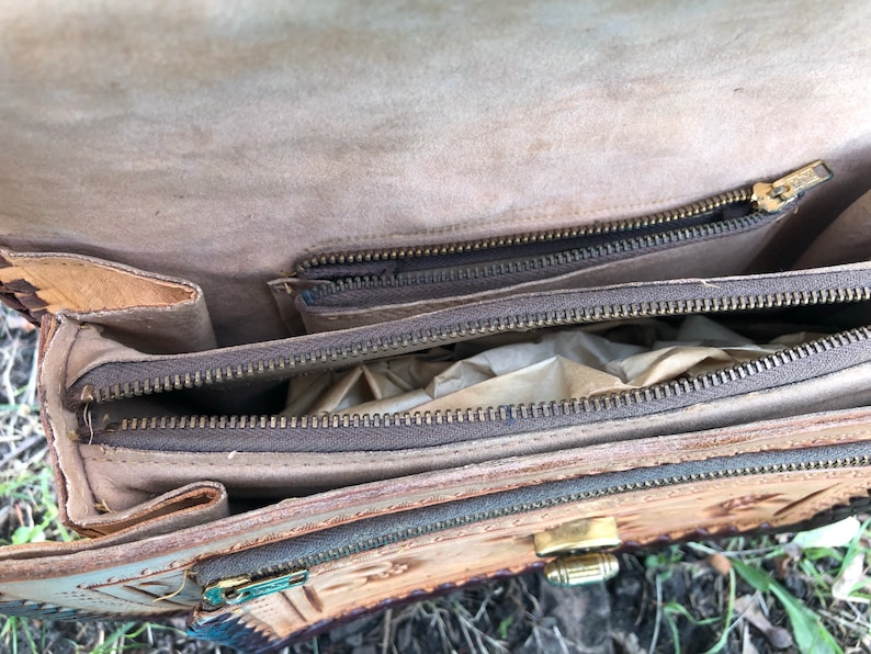 May include: Open view of a vintage leather satchel. The bag is tan with a detailed embossed design and multiple zippered compartments. The zippers and clasp are gold-toned. The interior is lined with a lighter tan material.