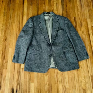 May include: A gray tweed blazer with a two-button closure, two front pockets, and a notched lapel. The blazer has a light gray lining and is laid out on a wooden floor.