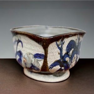 以下が含まれることがあります： 茶色の縁取りと青と白の花柄が施された、小さな白い陶器のボウル。ボウルはわずかに丸みを帯びた底をしています。