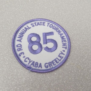 May include: A white and purple embroidered patch with the text "3rd Annual State Tournament - CYABA Greeley" and the number "85" in the center.
