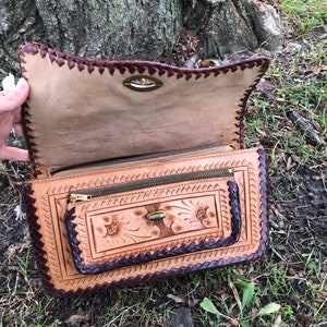 May include: Open, tan leather handbag with a decorative floral design and a dark brown border. The bag features a zipper pocket and a clasp closure. The edges are intricately stitched, and the bag is held open, revealing the interior.