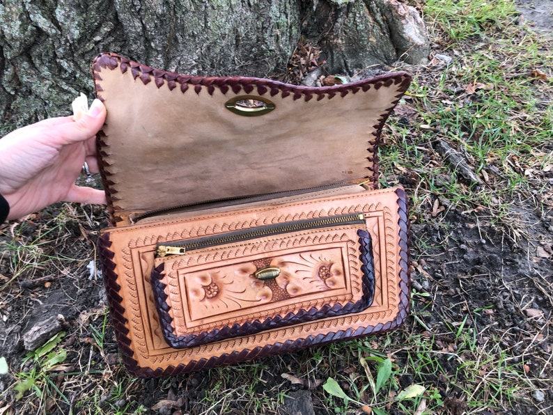 May include: Open, tan leather handbag with a decorative floral design and a dark brown border. The bag features a zipper pocket and a clasp closure. The edges are intricately stitched, and the bag is held open, revealing the interior.
