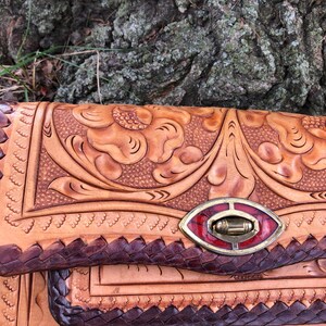 May include: A handcrafted leather clutch with intricate floral tooling. The bag features a braided edge, a decorative clasp with red accents, and a natural tan color with darker brown details. The design showcases detailed craftsmanship.
