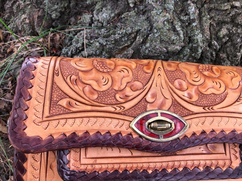 May include: A handcrafted leather clutch with intricate floral tooling. The bag features a braided edge, a decorative clasp with red accents, and a natural tan color with darker brown details. The design showcases detailed craftsmanship.