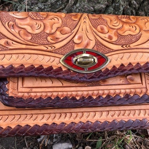 May include: A handcrafted leather clutch with intricate floral tooling. The natural tan leather is accented with dark brown braided edges and a decorative clasp with red and gold details. The bag's design showcases a blend of rustic charm and skilled craftsmanship.