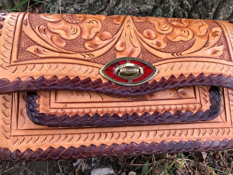 May include: A handcrafted leather clutch with intricate floral tooling. The natural tan leather is accented with dark brown braided edges and a decorative clasp with red and gold details. The bag's design showcases a blend of rustic charm and skilled craftsmanship.