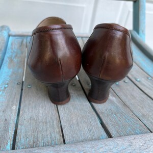 May include: A pair of brown leather heeled shoes with a pointed toe. The shoes are sitting on a wooden surface.