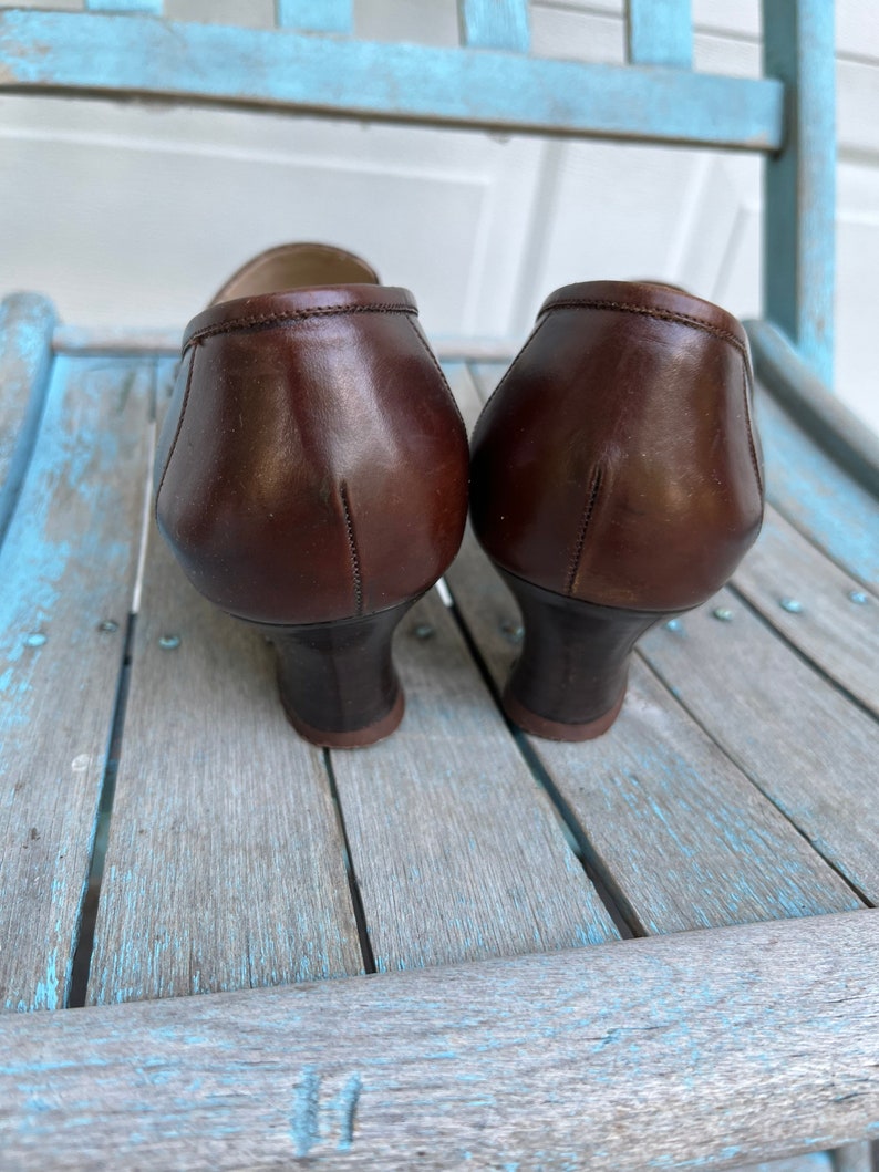 May include: A pair of brown leather heeled shoes with a pointed toe. The shoes are sitting on a wooden surface.