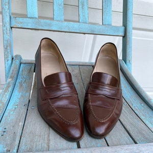 May include: A pair of brown leather loafers with a pointed toe and a single strap across the top. The loafers are sitting on a light blue wooden chair.