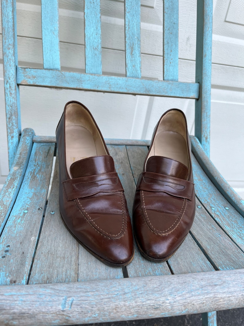May include: A pair of brown leather loafers with a pointed toe and a single strap across the top. The loafers are sitting on a light blue wooden chair.
