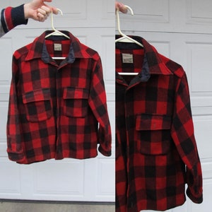 Peut inclure: Une chemise en flanelle à carreaux rouges et noirs avec deux poches avant et un col bleu foncé. La chemise est suspendue à un cintre blanc. La chemise est de style bûcheron classique.