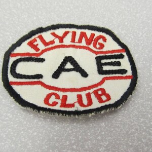 May include: White oval embroidered patch with black and red stitching. The patch says "Flying CAE Club".