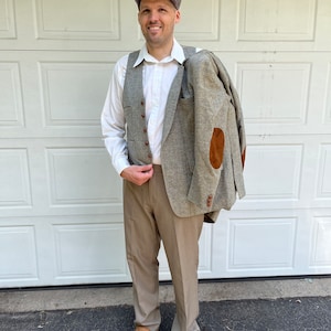 Puede incluir: Una chaqueta de tweed gris con coderas marrones se lleva sobre el hombro. El atuendo incluye un chaleco gris, una camisa blanca abotonada, pantalones color beige, una gorra gris y mocasines de ante marrones. El conjunto sugiere un estilo vintage o clásico.