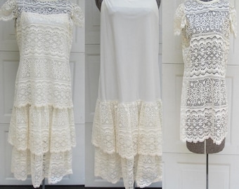 May include: Three vintage dresses on dress forms. The dresses are white and cream-colored, featuring lace and tiered designs. One dress has short sleeves and a lace overlay, another is a simple slip dress, and the third has a lace overlay and short sleeves.