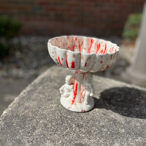 May include: A white ceramic bowl with red splatter paint design, featuring two cherub figurines on the base.