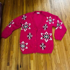 May include: A bright fuchsia long-sleeve cardigan sweater featuring a V-neckline and a front button closure. The knit garment is decorated with a repeating pattern of geometric snowflake-like shapes in contrasting white and black thread colors.