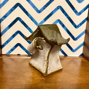 May include: A small, white ceramic house with a green roof. The house has a curved, scroll-like design on the side. The house is sitting on a wooden shelf.