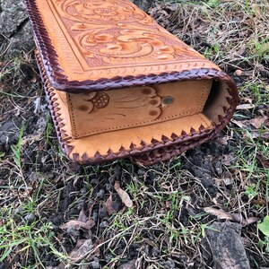 May include: A handcrafted, light brown leather box with intricate floral and scrollwork carvings. The edges are trimmed with dark brown leather and a decorative stitch. The box is rectangular and appears to be a storage container.