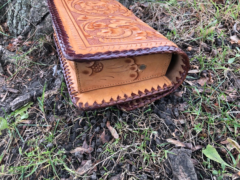 May include: A handcrafted, light brown leather box with intricate floral and scrollwork carvings. The edges are trimmed with dark brown leather and a decorative stitch. The box is rectangular and appears to be a storage container.
