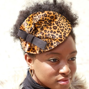 May include: A woman with curly black hair is wearing a black turtleneck shirt and a brown and black leopard print hat with a black bow. She is wearing gold triangle earrings.