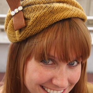May include: A woman with long, red hair is wearing a brown and gold knitted hat with a black net veil and a brown bow with rhinestones.