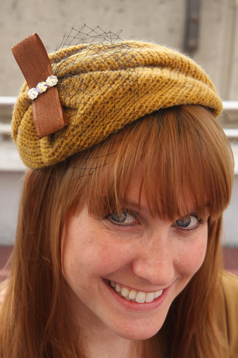 May include: A woman with long, red hair is wearing a brown and gold knitted hat with a black net veil and a brown bow with rhinestones.