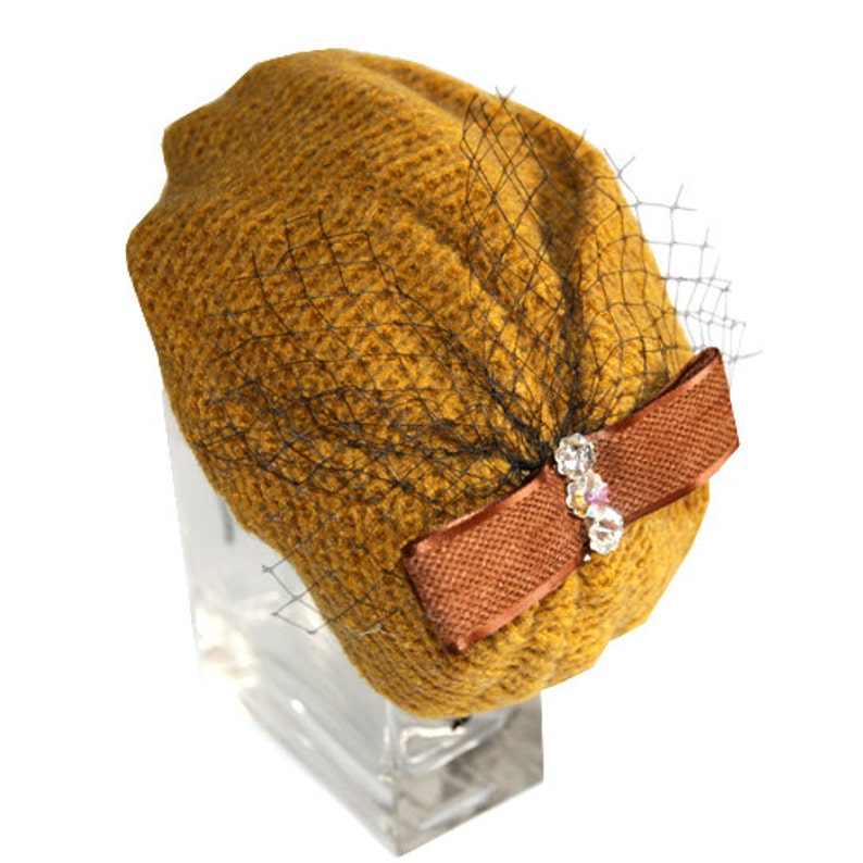 May include: A mustard yellow knitted beret with a brown velvet bow and a sheer black veil. The bow has a clear rhinestone embellishment.