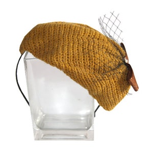 May include: A mustard yellow knitted beret with a black mesh veil and a brown bow.