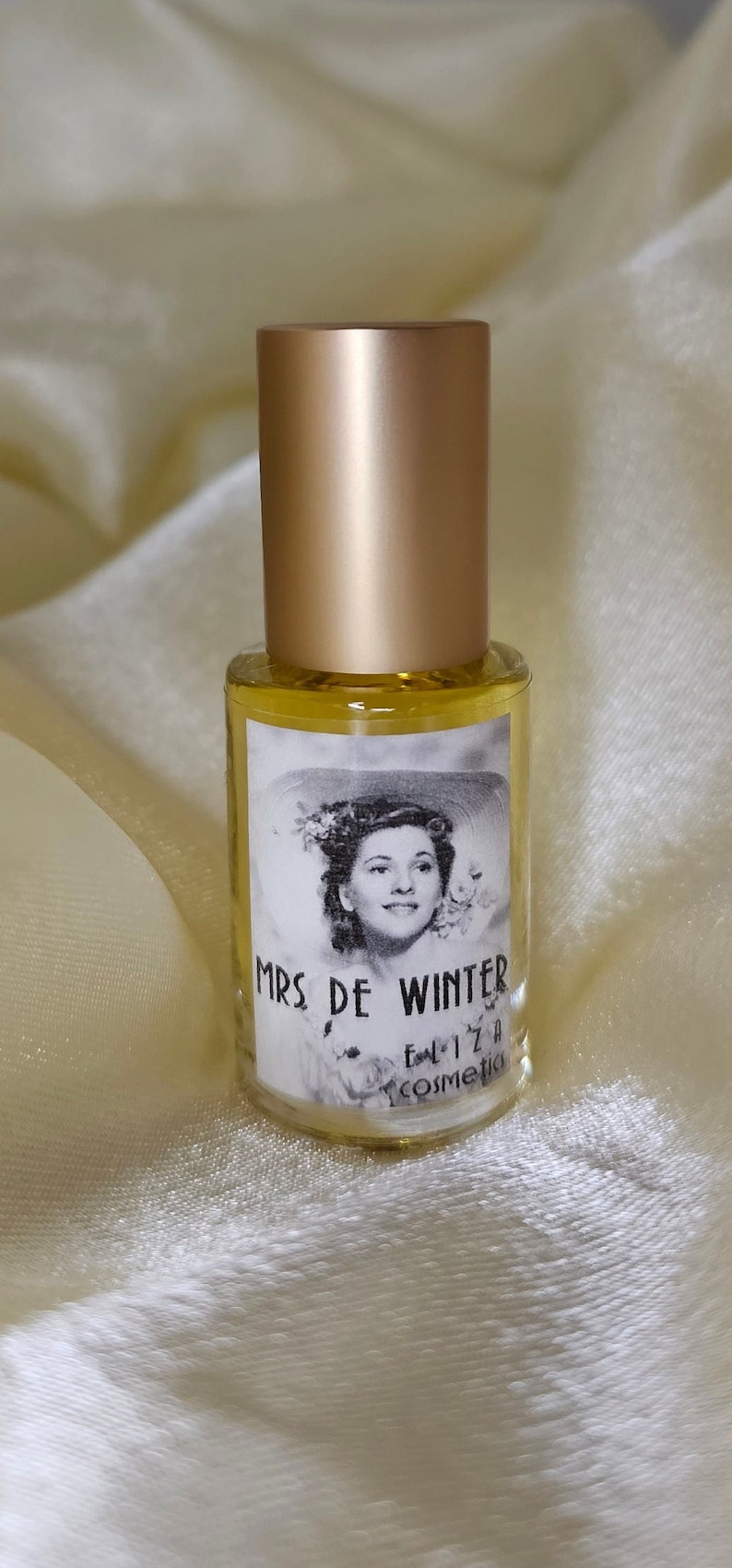 May include: A gold bottle with a clear label that says "MRS DE WINTER" and "Eliza Cosmetics". The label also features a black and white image of a woman wearing a hat.